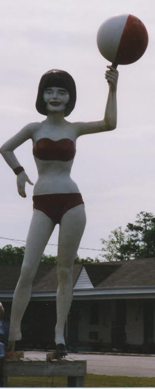 An 18 foot tall statue of a dark-haired woman wearing a red bikini and heels holding a red and white beach ball up in the air with her left hand. Behind her is an old motel.