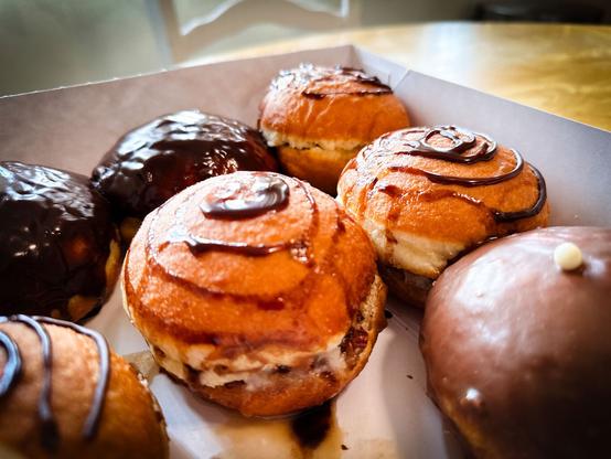A box of assorted pączki, featuring chocolate-glazed and cream-filled buns with chocolate drizzles on top. The pastries have a golden-brown appearance and are arranged closely together.