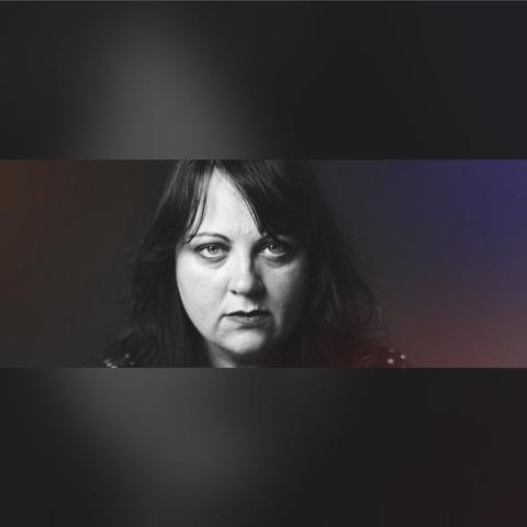 A black-and-white portrait of a woman with dark hair, staring directly at the camera with an intense and serious expression. Her face is softly lit, contrasting against a dark, moody background.