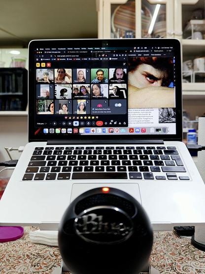 Una laptop MacBook abierta sobre una base y detrás de un micrófono esférico negro. En la pantalla, entre ventanas de imágenes y texto, una sesión de Google Meet, donde varias personas sonríen a la cámara desde diferentes lugares. Una de ellas toma la foto con un celular.