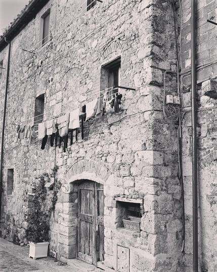 A black-and-white photograph of an old stone building with a weathered wooden door, small windows, and laundry hanging from a line. The rough texture of the walls and the simple everyday scene evoke a timeless, rustic charm.