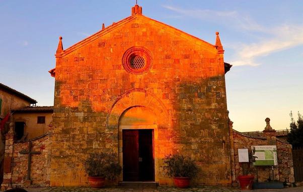 A historic stone church bathed in golden sunlight, with its entrance slightly ajar. The warm tones of the setting sun highlight the textured facade, creating a striking contrast against the clear blue sky.
