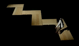 A still from the By Storm video with a black background, wooden zig zagging panels, and rapper RiTchie walking on top of them