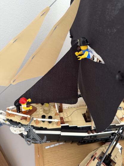 A close-up view of a LEGO pirate ship, featuring two mini-figures: one with a red hat at the helm and another in a black hat working on the sails. The ship has two sails—one beige and one black—set against