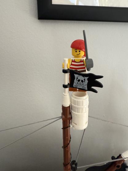 A Lego pirate figure with a red hat and striped shirt stands atop a ship's mast, holding a sword and a black flag featuring a skull and crossbones. The background is a plain wall, and a framed picture can be seen above the mast.