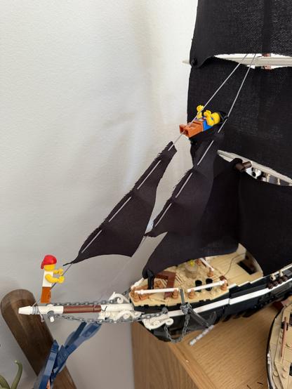 A model ship with two LEGO figures, one at the bow holding a rope and another hanging from the mast. The ship features black sails and is displayed on a wooden shelf against a light-colored wall.