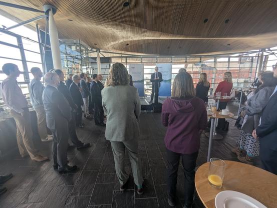 A small crowd of people gathers in the Oriel at the Senedd in Cardiff Bay to listen to someone in a suit speaking at a lecturn.