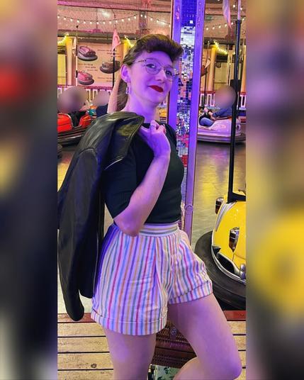 Photography of a woman wearing a retro fashioned outfit with colourful striped shorts and a dark knit jumper leaning against the post of an old fashioned bumper car fair atttraction. The woman is slightly smiling into the camera, wearing glasses and her hair styled into a high ponytail accentueted with a scarf tied into a bow and a bumper roll as fake bangs.
The whole surrounding is rather dark and lit with lights in different colours.