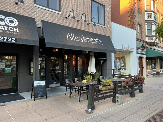 Outdoor view of Alfio's Buon Cibo, an Italian-Argentine restaurant with a patio, black awning, and neighboring shops on a city street.