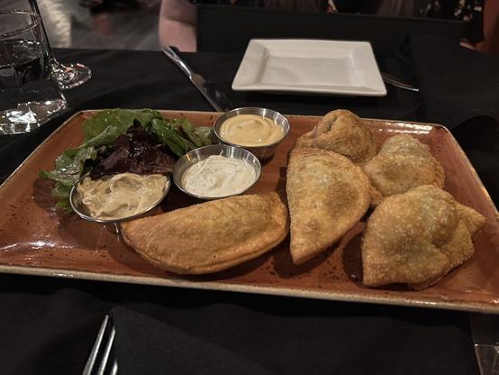 A plate of golden-brown fried empanadas with three dipping sauces and mixed greens, served on a rustic dish at a restaurant.