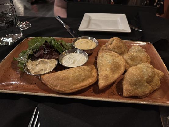 A plate of golden-brown fried empanadas with three dipping sauces and mixed greens, served on a rustic dish at a restaurant.