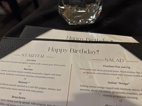 A restaurant menu with "Happy Birthday!!" at the top, featuring starters and salads, placed on a black tablecloth with a glass nearby.