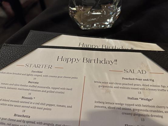 A restaurant menu with "Happy Birthday!!" at the top, featuring starters and salads, placed on a black tablecloth with a glass nearby.