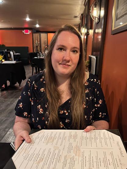 A woman with long brown hair and a floral dress sits in a warmly lit restaurant, holding a menu with "Happy Birthday!!" printed on it.