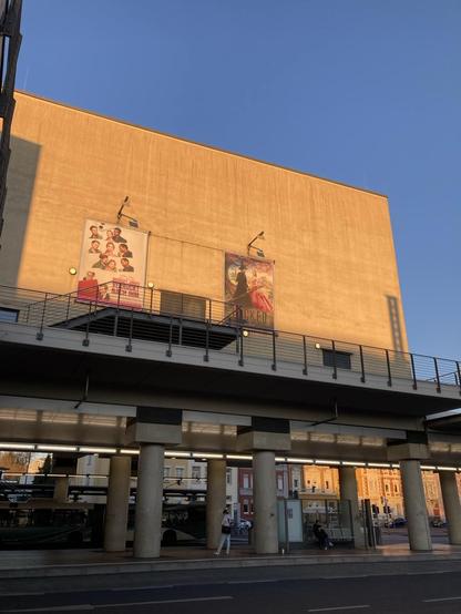 Foto zeigt eine von der Abendsonne angestrahlte Wand, deren fader gelber Anstrich nun orange leuchtet. Darauf sind zwei Plakate Kinowerbung platziert. Darunter befindet sich der Busbahnhof. Zu sehen ist ein Geländer und darunter die Betonsäulen. Auch ein Bus, aber alles liegt im SchattenY Oberhalb des Gebäudes ist der Himmel ganz in blauem Ton zu sehen. Das nun liegt auch an der Handykamera, die gerne übertreibt und den Himmel  blauer darstellt als er tatsächlich war.