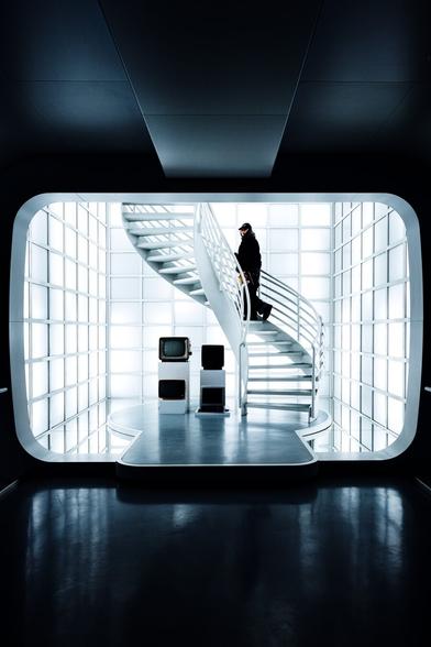 A futuristic, minimalistic interior with a white spiral staircase leading to an upper level. The walls consist of illuminated glass panels, casting a cool, soft glow throughout the space. A person dressed in black, wearing a cap, descends the staircase, adding a contrasting human element. In the foreground, a dark reflective floor enhances the depth of the image. To the left of the staircase, a small display of vintage televisions sits on white pedestals, juxtaposing retro technology against the sleek, modern environment. The scene has a cinematic, sci-fi atmosphere with a strong sense of symmetry and composition.
