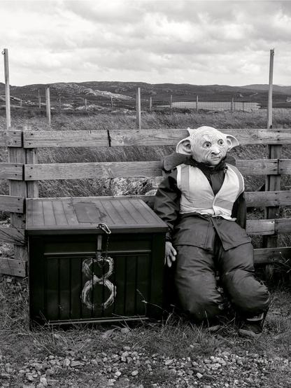 A funny man with pointy ears is sitting against a wooden fence next to a padlocked trunk. In the background are mountains and a typical Scottish lake.
