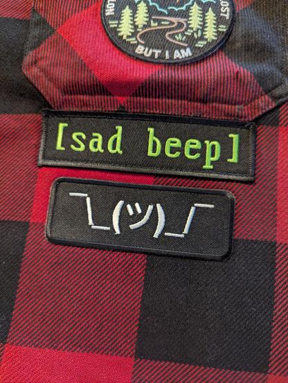 A shrugging text emoji patch placed just under a "[sad beep]" patch which itself is under a pocket on a buffalo check coat.