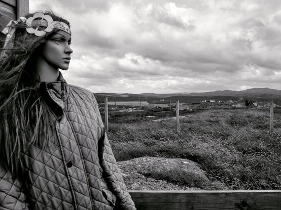 A female plastic mannequin wearing a quilted jacket and bandana decorated with a flower brooch is leaning against a wooden wall. She seems to be looking out at the horizon. In the background are houses, mountains and a typical Scottish lake.