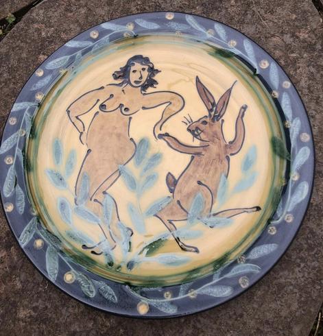 Round ceramic plate decorated with a dancing woman and March Hare using slip-trailing and painting.