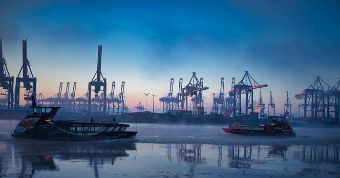 Two public transport ferries cross the icy waters of the Hamburg harbor on a winter morning, possibly during a shift change. Workers, perhaps from Airbus or the docks, head home from the night shift while others start their day. The sky glows with the soft hues of dawn, while towering cranes of the container terminal loom in the misty background, standing as symbols of the city’s maritime industry.