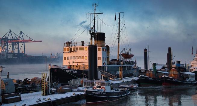 The historic icebreaker Stettin moored among other vintage ships in the Museum-Port of Hamburg Övelgönne on a winter morning. The sunrise casts a soft golden glow on the snow-dusted decks and the calm harbor water, while mist rises in the background, blending with the silhouettes of industrial cranes. A moment where maritime history meets the present.