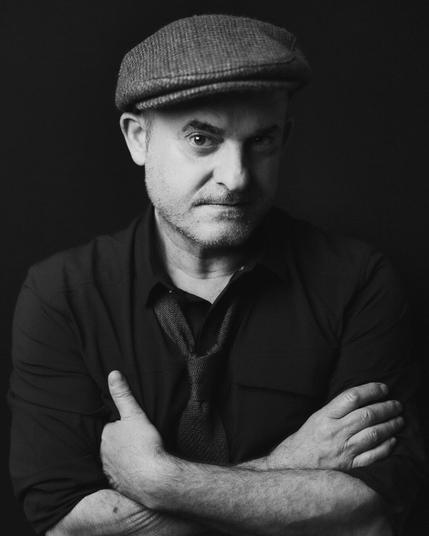 A striking black-and-white portrait of Roberto, wearing a flat cap and dark button-up shirt with a loosely knotted tie. His arms are crossed, and he looks directly into the camera with quiet intensity, exuding a mix of confidence and mystery. The moody lighting enhances the depth of his expression.