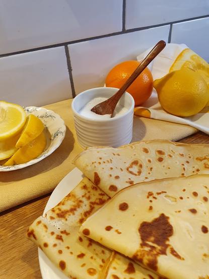 A plate of golden-brown pancakes is displayed on a wooden table, accompanied by a small dish of sugar featuring a wooden spoon. Nearby, slices of lemon and a whole lemon sit alongside an orange, with a partially visible cloth in the background.