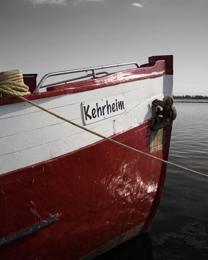 A fishing boat named Kehrheim moored on Fehmarn Island. The hull is red and white, contrasting against a desaturated background of sky and water. The boat's rope stretches diagonally, leading toward the calm sea. The selective color editing draws attention to the vessel while maintaining a timeless and artistic feel.