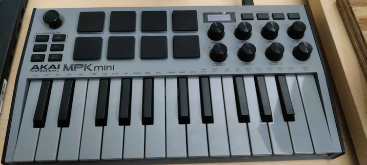 A Picture of a gray Akai MPK Mini mk3 Midi controller, it has two octaves worth of piano keys, 8 knobs and 8 drum pads as well as a small joystick for controlling instruments settings, etc