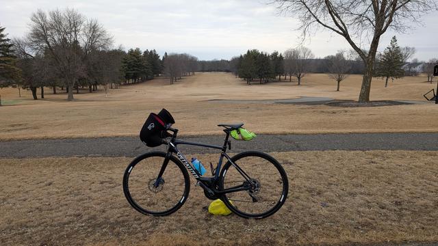 Blue and black Roubaix bicycle parked off the Eastwood Trail with the 3rd hole of the Eastwood Golf Course behind. The fairway is brown, there are bare trees and evergreen trees lining the fairway, and the brown "green" is in the distance under cloudy skies.