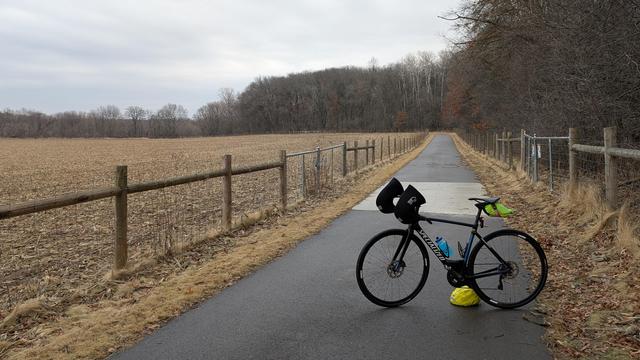 Blue and black Roubaix bicycle parked on the damp asphalt Chester Woods State Trail which goes straight behind before curving to the left. There are wooden posts holding wire fencing on both sides of the trail, deep, bare woods on the right and curving ahead in the distance to the left, and a brown stover covered farm field on the left under cloudy skies.