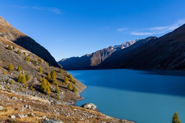 Ein türkisfarbener Stausee an einem Herbstmorgen in den ersten Sonnenstrahlen. Ein paar Lärchen zieren die felsige Umgebung.