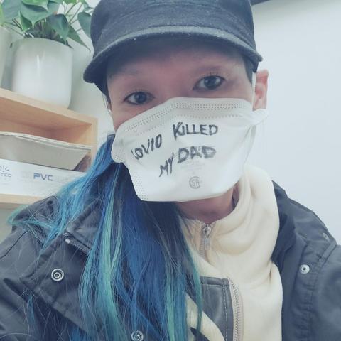 Me, wearing a white medical mask with handwritten text in black marker that reads "COVID KILLED MY DAD." Wearing a black jacket over a light-colored hoodie. A plant & boxes are visible in the background.