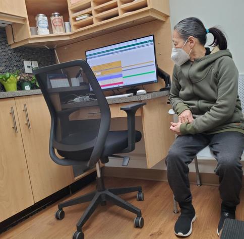 Mom, wearing an N95 mask sitting in doctor's office waiting to be seen. She is wearing a green hooded sweatshirt & grey sweatpants. She is sitting next to a desk with a computer displaying a medical schedule. Various medical supplies such as bandages & cotton are displayed on shelves above the desk. The office has a clinical yet somewhat cozy feel with potted plants on the counter.