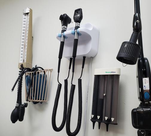 Various medical examination tools are mounted on a white wall in a doctor's office, including a mercury sphygmomanometer, otoscope & ophthalmoscope, Welch Allyn dispenser & articulating examination light.