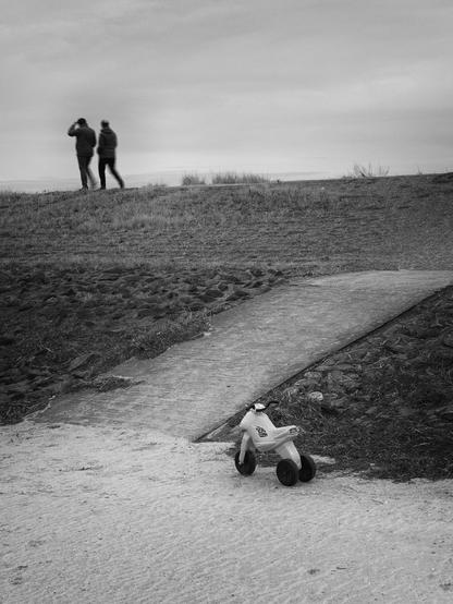 The black-and-white photograph depicts a gently sloping path leading from a sandy area in the foreground up to a small hill. At the lower edge of the path, a small plastic children's tricycle stands abandoned. It appears unused, as if left behind.

In the upper left part of the image, at the highest point of the path, two people can be seen walking away from the viewer. They are slightly blurred, caught mid-step, seemingly engaged in conversation. The sky is mostly overcast, with soft, diffused light. The surrounding landscape is sparse, with patches of grass and low vegetation lining the path.

The composition is divided into three layers: the sandy foreground with the toy, the rising path, and the sky with the two distant figures.