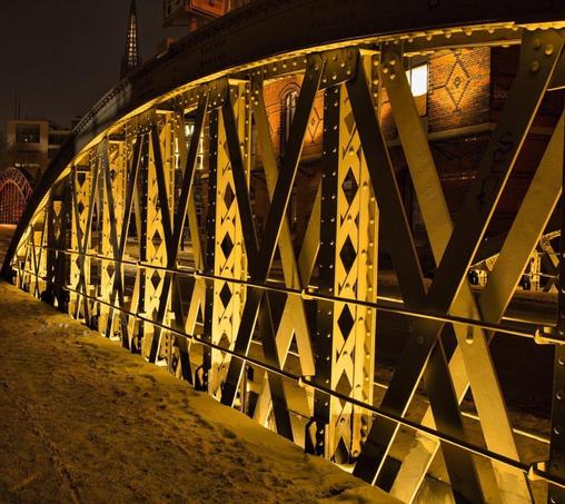 A detailed close-up of an iron bridge in Hamburg's Speicherstadt, illuminated in warm golden light. The intricate metal framework stands out against the dark night sky, emphasizing the industrial elegance of the city's historic port district.
