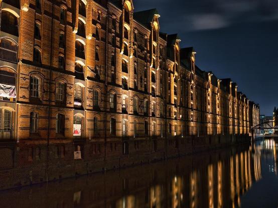 A mesmerizing shot of the Speicherstadt warehouses at night, their historic facades bathed in golden light. The still water below perfectly mirrors the illuminated buildings, creating a breathtaking play of architecture and reflection.