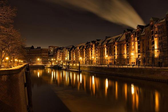 A stunning nighttime view of the Speicherstadt in Hamburg, with historic red-brick warehouses reflecting on the calm canal waters. The golden glow of streetlights and the soft trails of smoke rising into the night sky create a dreamy, timeless atmosphere.