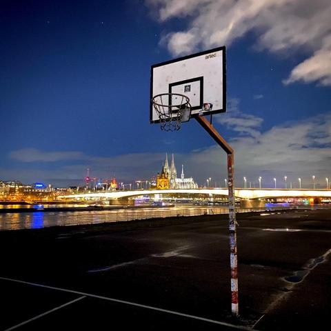 A picture by Alex depicting a basketball basket (however it's called) and, behind, the Kölner Dom (Cologne's cathedral) and the Rhine river, after sunset, getting dark, with city lights.