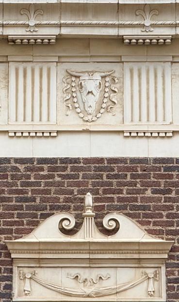 A dentillated course carved in light tan stone above a brick wall. The most prominent feature is a longhorn cattle skull in bas relief. Below is an inverted ionic cap on a door.