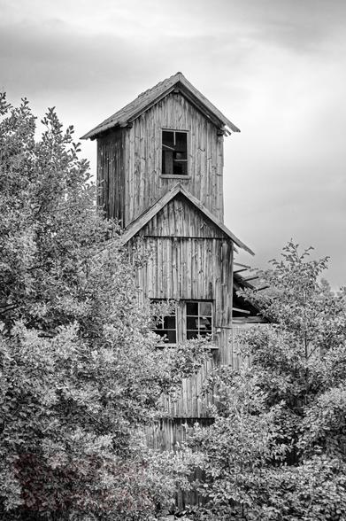 Schwarzweißfotografie im Format 3:2 waagerecht.
Ein mehrstöckiges turmartiges Gebäude in Holzkonstruktion und mit Holzbrettern verkleidet. Eingezwängt in Bäume und Büsche. Im Hintergrund, Unwetter ankündigende Wolken.