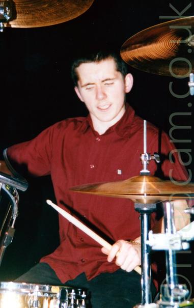 Close-up of a man with short dark hair, wearing a red shirt and black pants, playing a set of drums.
Gold-coloured cymbals seem to hang in midair in front of him.
The wall behind him is black.