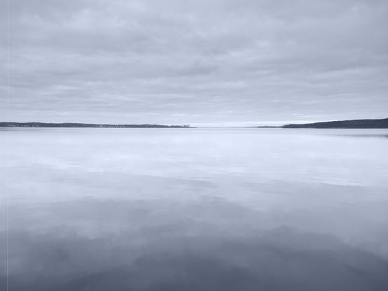 The image shows a peaceful, expansive body of water under a cloudy sky. The water is calm, creating a mirror-like reflection of the sky above. In the distance, there is a low horizon line with land masses visible. The image is in black and white, which gives it a serene and timeless quality.