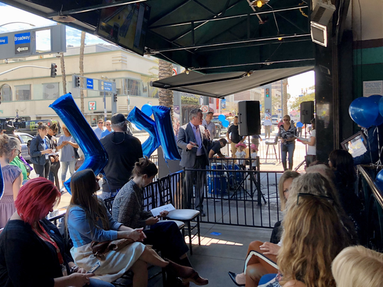 People sitting and standing outside on a sidewalk with balloon letters spelling the call letters of the radio station. Someone is speaking into a microphone