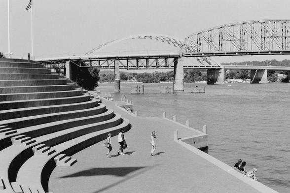 Black and white image of a large car bridge over a river with a winding wall of stepped concrete running along one side of the river. A few people sit on the edge of the river and three young men walk towards the river's edge wearing shorts and t-shirts. One of them has a large boombox cassette deck with a large strap draped over his shoulder.