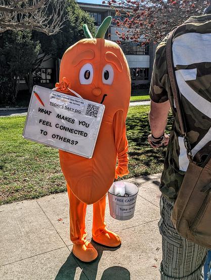 Carrot mascot carrying a bucket of carrots and sign reading "what makes you feel connected to others."