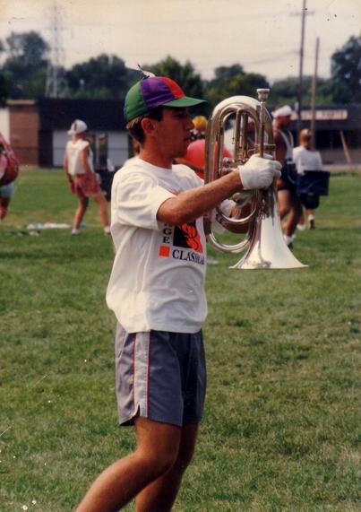 A young man (me, not even 20 years old yet) wearing a colorful cap with a propeller and a white t-shirt, holding a baritone bugle while marching on a grassy field. I am in motion, surrounded by other people, my fellow Glassmen, engaged in a drum corps practice.