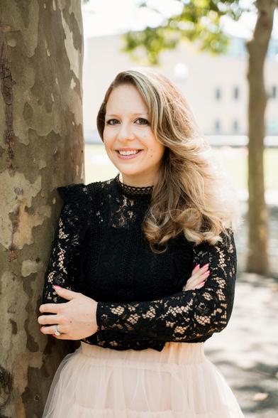 Head-and-shoulders color photo of Daria Lavelle, a fair-skinned woman with dark eyes and long, ash-blonde hair. She wears a black lace top with a high collar and a pink tulle skirt. She stands outside, near a stone wall. With her face in three-quarter view, she looks directly into the camera and beams sunnily.